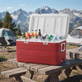 A red cooler filled with iced beverages sits on a wooden picnic table outdoors at a campsite, with tents, trees, and snow-capped mountains in the background.