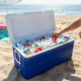 A blue and white cooler filled with ice and bottled drinks sits open on a sandy beach, with a person's arm reaching in to grab a red can. The ocean and beach umbrellas are visible in the background.
