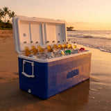 A blue and white cooler on a sandy beach at sunset, filled with ice and glass beer bottles. The cooler has a visible 'Tide' logo on the front side.