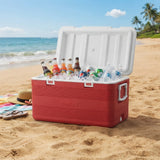 A red and white cooler filled with ice and assorted bottled drinks sits open on a sandy beach, with ocean waves, blue sky, and palm trees in the background.