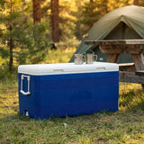 A blue and white cooler sitting on grass in a campsite setting, with a green tent and wooden picnic table blurred in the background. Two metal mugs rest on top of the cooler.